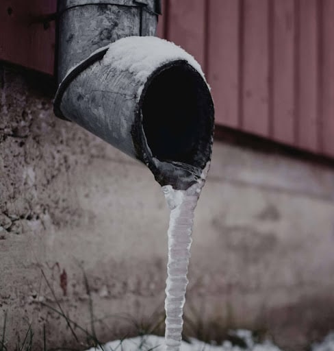 A metal drainpipe with a long icicle hanging from it, partially covered in snow, against a textured wall.