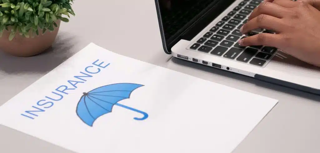 Man typing on a laptop with a piece of paper with the word 'insurance' typed on it and a blue umbrella underneath the word.